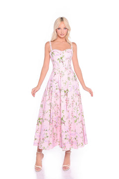 A woman wears the Rosella Blush Pink Floral & Bow Print Casual Dress, featuring a fitted bodice and flared skirt—perfect for summer weddings—paired with strappy heels, standing against a white background.