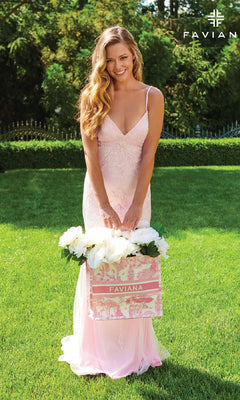 A woman in a Strappy-Back Long Light Pink Prom Dress by Faviana stands on grass, holding a pink and white Faviana bag filled with white flowers. Trees and shrubs are in the background, with the Faviana logo in the top right corner.