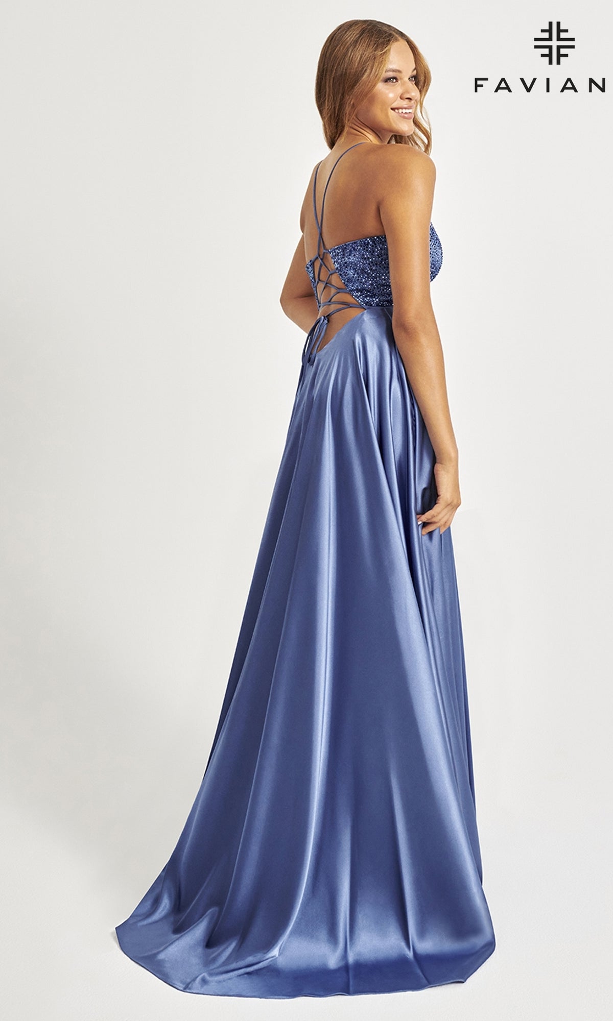 A woman in the Faviana Beaded-Bodice Long Prom Dress S10870, featuring blue satin, a beaded v-neck bodice with thin straps, an a-line silhouette, lace-up back, fitted bodice, and flowing skirt, stands facing away looking over her shoulder.