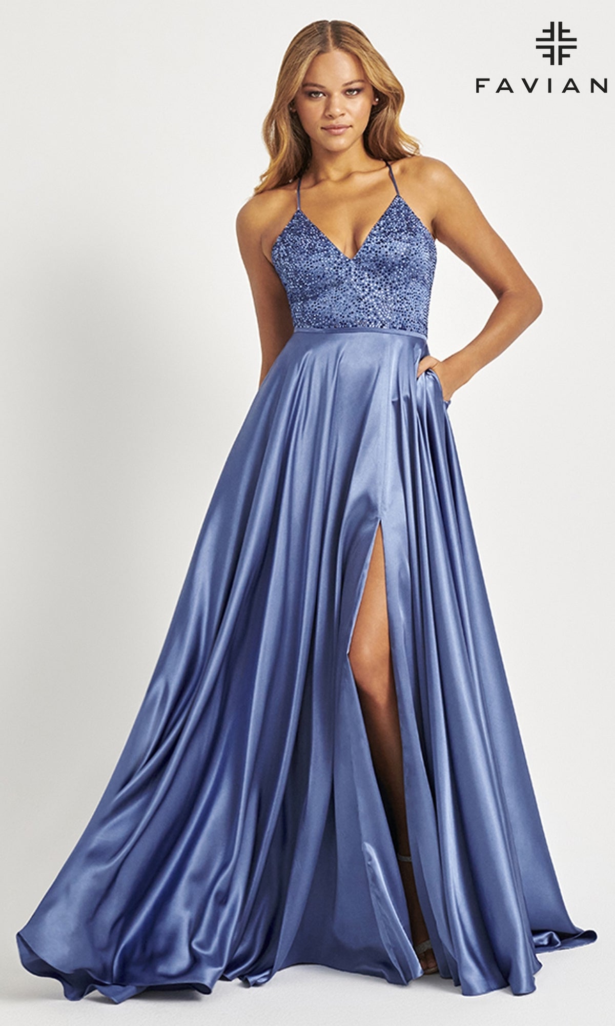 A woman wears the Faviana Beaded-Bodice Long Prom Dress S10870, featuring a sleeveless blue satin design with thin straps, a beaded V-neck bodice, floor-length skirt, and high slit, standing against a plain background.