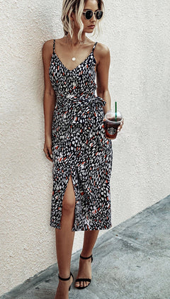 A woman in a V-Neck Casual Print Midi Wrap Dress with Front Slit stands against a light wall, wearing sunglasses, holding an iced drink, and gazing to the side.