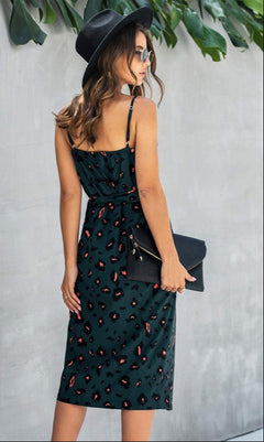 Woman stands outdoors under green leaves, wearing sunglasses, a black hat, and the V-Neck Casual Print Midi Wrap Dress with Front Slit in a red and tan spotted pattern, holding a black clutch bag.