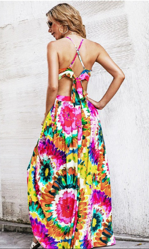 A woman stands against a textured white wall wearing the Casual Open-Back Colorful Print Maxi Dress, featuring crisscross adjustable spaghetti straps and a vibrant print.