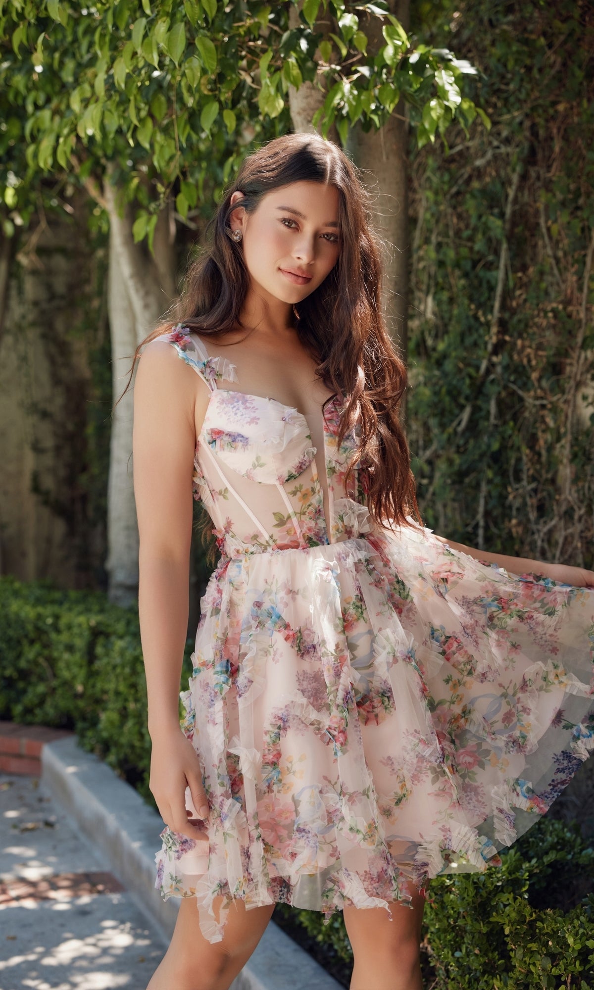 Wearing the Short Pink Floral-Print Homecoming Dress S849, a woman stands outdoors before lush greenery, gracefully holding one side of her skirt to showcase the corset-style bodice and floral-print design.