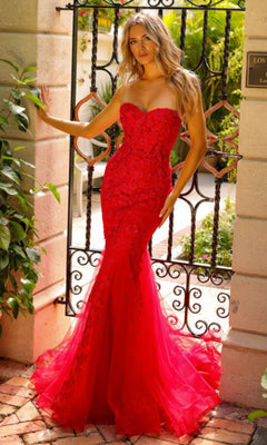 A woman stands outdoors before a wrought iron gate, wearing the Amelia Couture Corset Mermaid Prom Dress SU081—a strapless, form-fitting red gown with a flared tulle hem and delicate floral embellishments.