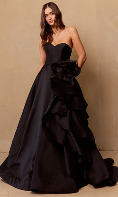 Woman wearing the Amelia Couture SU085 Strapless Prom Ball Gown, a black dress with a fitted bodice and full skirt adorned with dramatic ruffled detailing, standing against a beige wall.