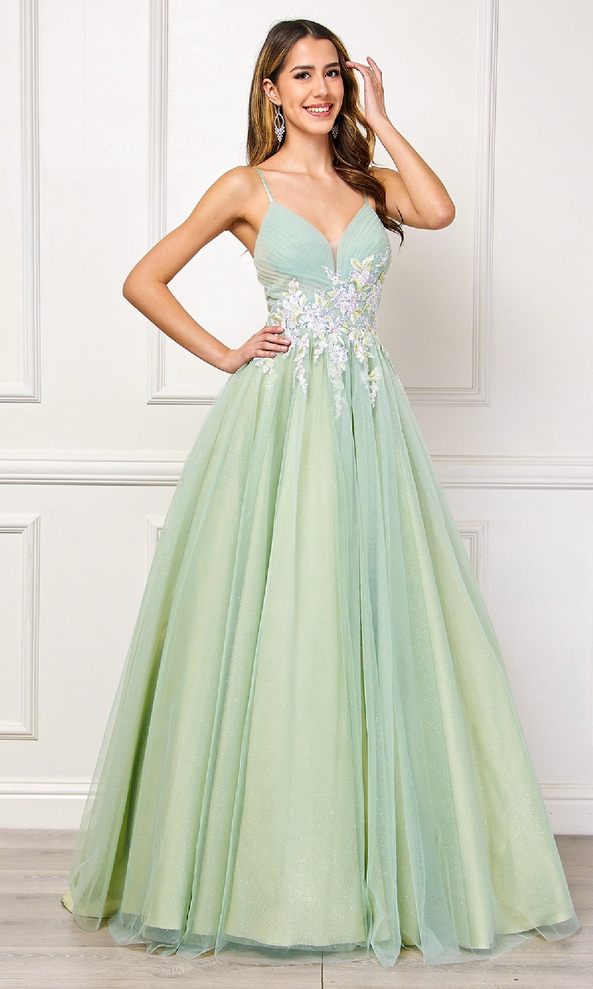 A woman stands indoors against a white paneled wall, wearing the Amelia Couture SU088 Long Pastel Prom Ball Gown, an a-line formal dress with floral appliqué details.