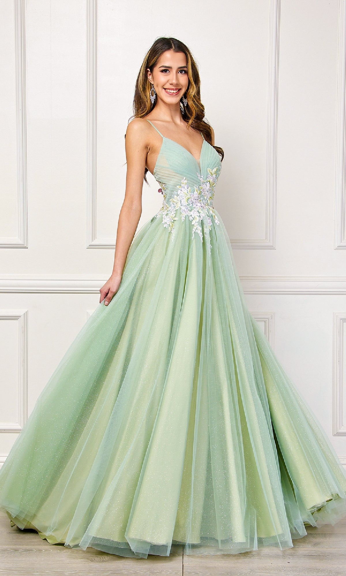 A woman poses with a smile and hand on her hip, wearing the Amelia Couture SU088 Long Pastel Prom Ball Gown—a light green, A-line formal dress with floral embroidery at the waist—against a white paneled wall.