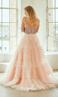 Woman modeling the back of a light pink ball gown with a lace-up corset back and tiered ruffle skirt in front of large windows.