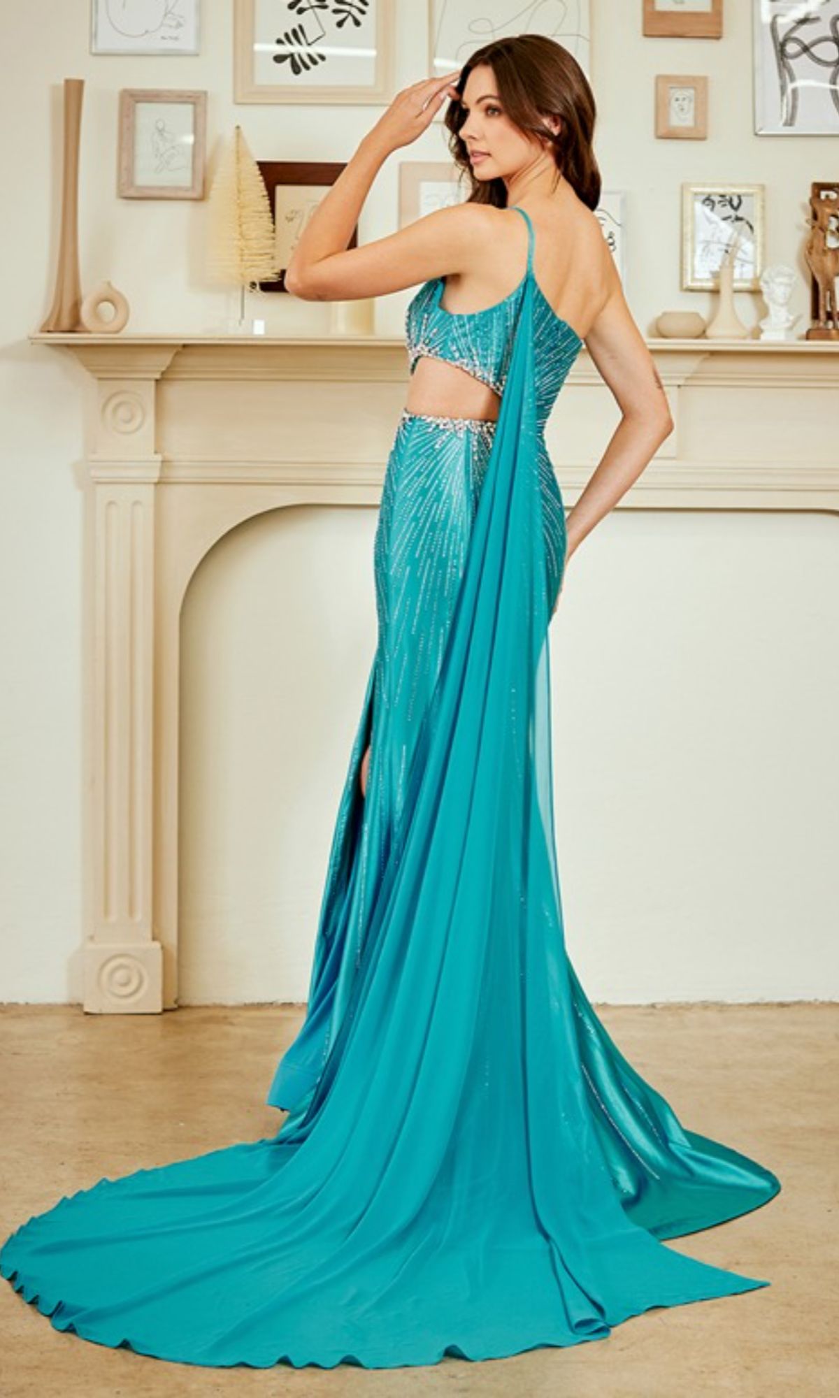 A woman stands indoors in front of a decorated mantel, wearing the Amelia Couture SU097—a turquoise long prom dress featuring a one-shoulder neckline and long train—looking over her shoulder.