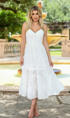 A woman stands outdoors by a stone fountain and greenery, looking at the camera in the Velvi Tea-Length White Graduation Dress Sisi—a sleeveless, button-down eyelet lace dress that adds elegance to her look.