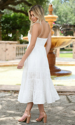 A woman in the Velvi Tea-Length White Graduation Dress Sisi and nude heels stands outdoors by a stone fountain, looking over her shoulder.