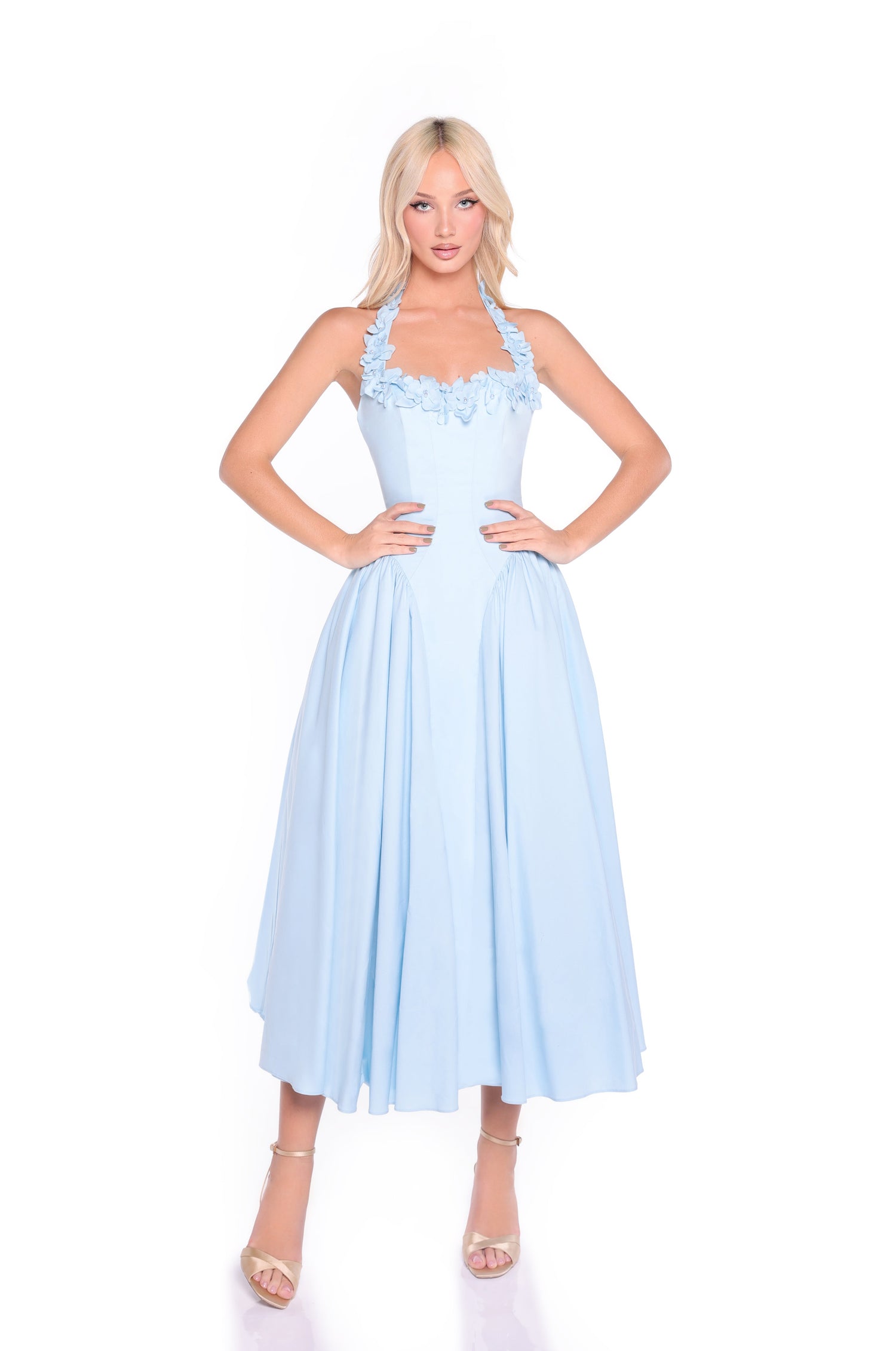 A woman stands with hands on hips, wearing the Skye Powder Blue Midi Casual Halter Dress featuring a full skirt and floral details, paired with strappy heels against a white background.