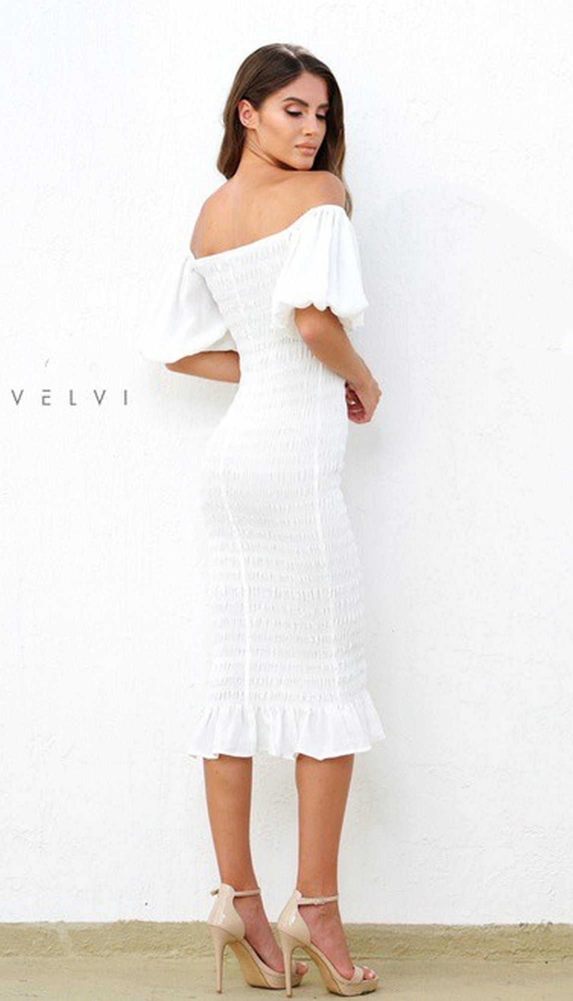 A woman stands against a white wall in the Skyla Knee-Length Puff-Sleeve Wedding-Guest Dress—a chic white off-the-shoulder style with puff sleeves and ruffled hem, paired with nude heels for a stylish event look.