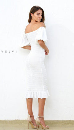 A woman stands against a white wall in the Skyla Knee-Length Puff-Sleeve Wedding-Guest Dress—a chic white off-the-shoulder style with puff sleeves and ruffled hem, paired with nude heels for a stylish event look.