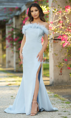 A woman in the Velvi Strapless Long Feather Prom Dress Sochi, a light blue gown with feather trim and a high slit, stands outdoors by pink flowers and stone columns.