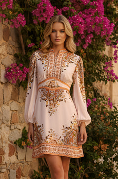 A woman in an ivory paisley print short casual dress, Sofia, stands before a stone wall covered with pink bougainvillea flowers.