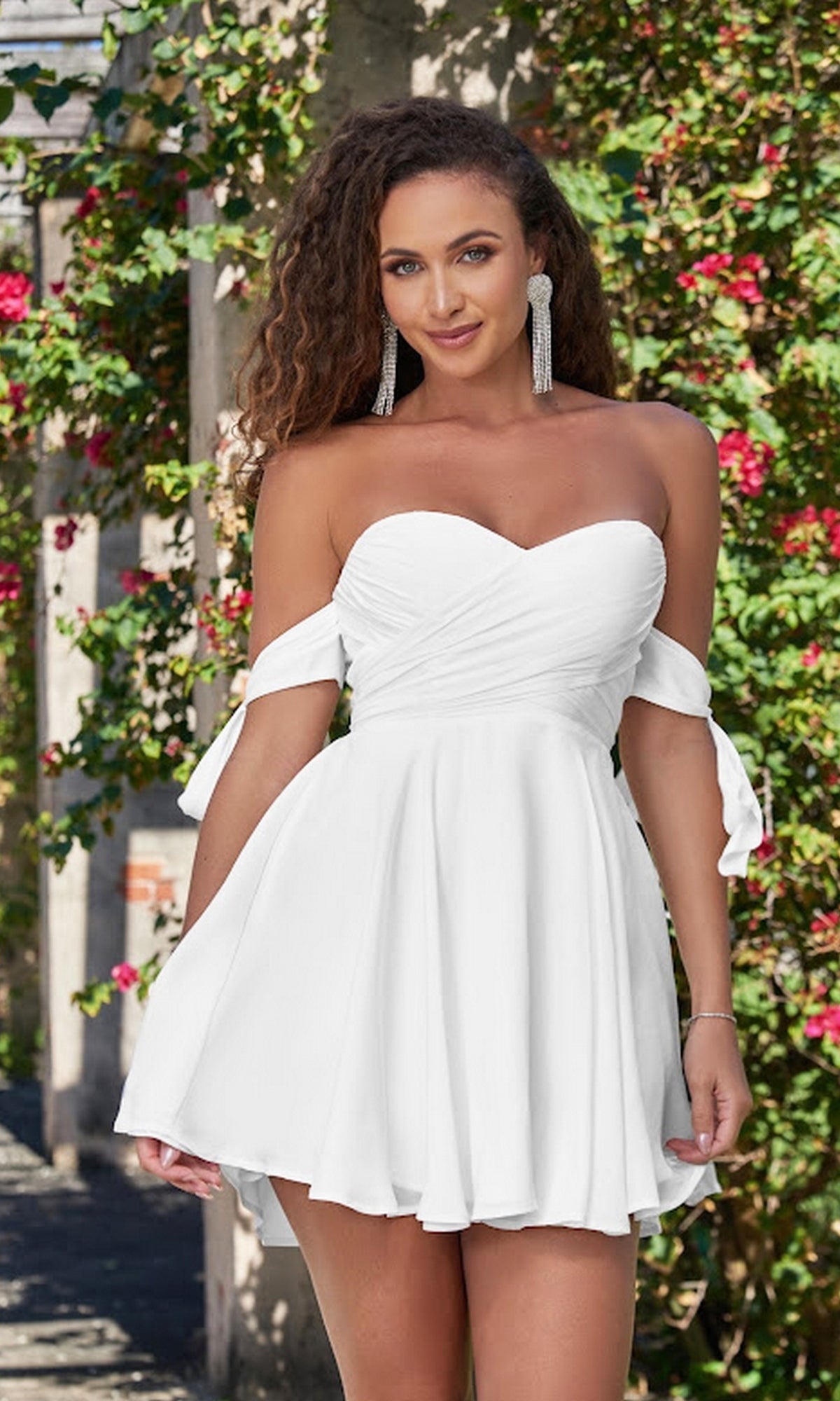 A woman stands outdoors before a flowering trellis, wearing the Velvi Off-the-Shoulder Mini Party Dress Sophie, a white mini dress featuring tie sleeves.