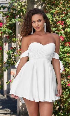 A woman stands outdoors before a flowering trellis, wearing the Velvi Off-the-Shoulder Mini Party Dress Sophie, a white mini dress featuring tie sleeves.