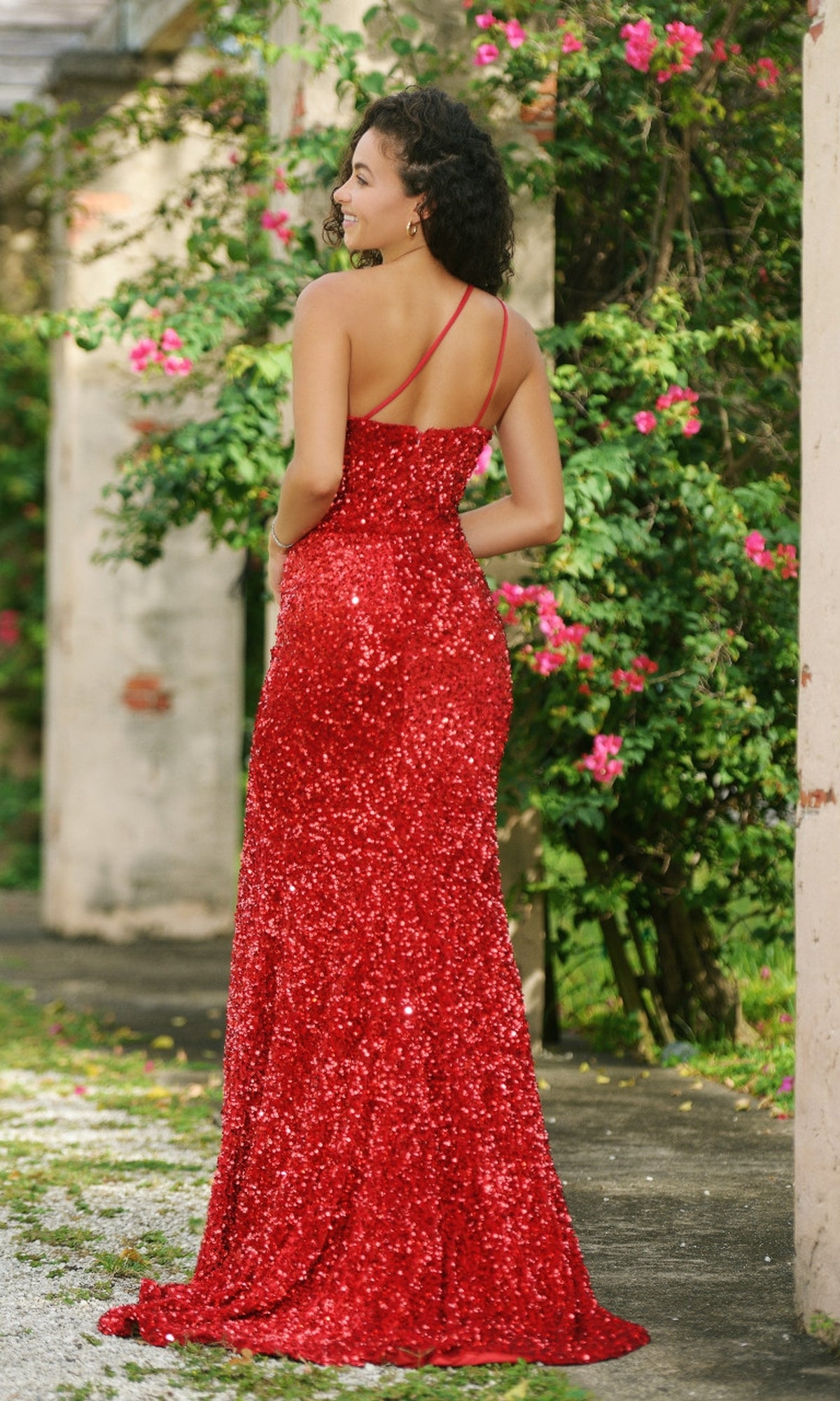 A woman in the Velvi One-Shoulder Long Sequin Prom Dress Stardust stands outdoors with her back to the camera, surrounded by greenery and pink flowers.