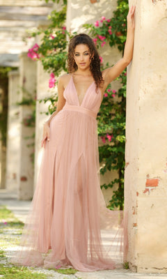 Wearing the Velvi Plunging V-Neck Long Tulle Prom Dress Sydney, a woman stands by a garden column in an ethereal light pink gown, its flowing tulle beautifully complemented by blooming flowers behind her.