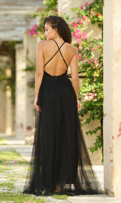 Outdoors, a woman with curly hair poses among columns and pink flowers in the Velvi Plunging V-Neck Long Tulle Prom Dress Sydney—a black, floor-length designer gown with a backless design and thin crisscross straps.