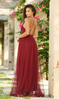 Outdoors, a woman wears the Velvi Plunging V-Neck Long Tulle Prom Dress Sydney—a sleeveless, floor-length burgundy gown with a strappy open back—framed by pink flowers and greenery.