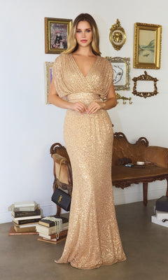 A woman wears the Cap-Sleeve Long Sequin Formal Dress: Amoris T101 in gold, standing in a stylish room with vintage decor, framed art, a brown sofa, and stacked books.