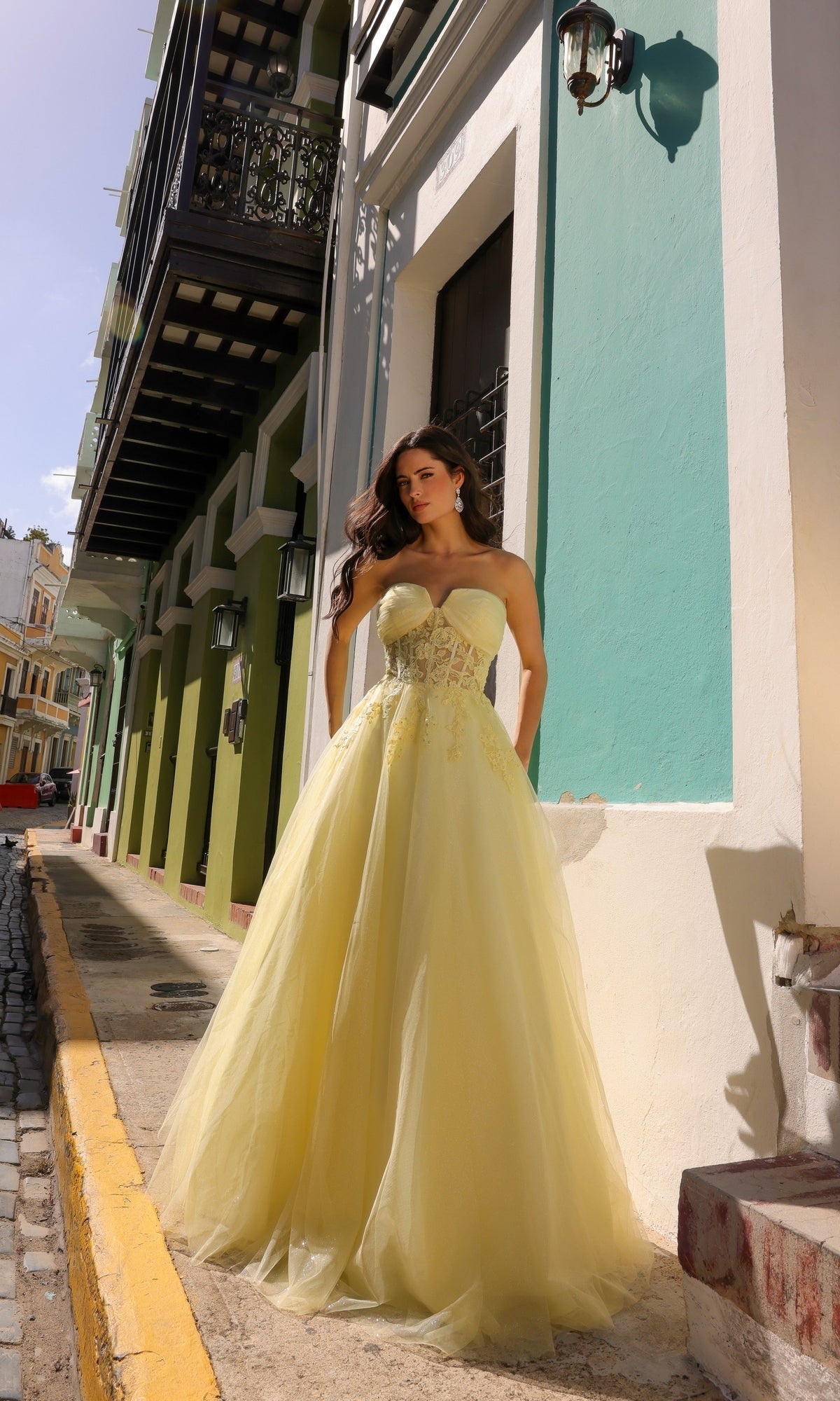 A woman in the Strapless Sheer-Waist Long Prom Ball Gown T1326 stands on a sunlit street next to colorful buildings with decorative balconies and lanterns.