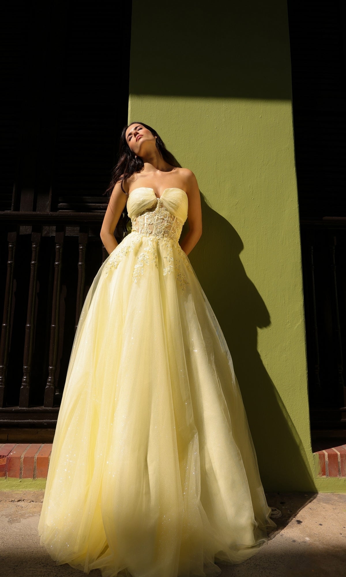 A woman wearing the Strapless Sheer-Waist Long Prom Ball Gown T1326 stands against a green wall, sunlight casting shadows behind her.