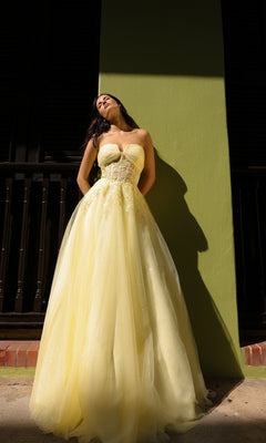 A woman wearing the Strapless Sheer-Waist Long Prom Ball Gown T1326 stands against a green wall, sunlight casting shadows behind her.