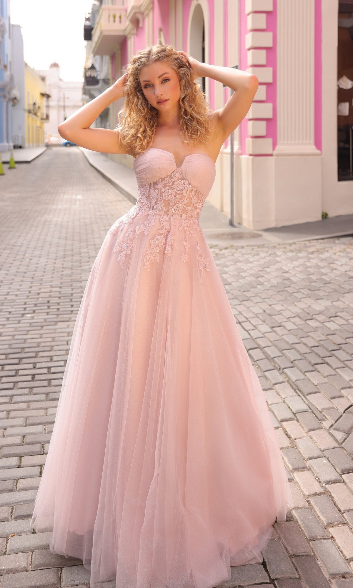A woman wearing the Strapless Sheer-Waist Long Prom Ball Gown T1326, featuring light pink tulle and floral embroidery, stands on a cobblestone street lined with pastel-colored buildings.