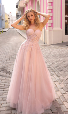 A woman wearing the Strapless Sheer-Waist Long Prom Ball Gown T1326, featuring light pink tulle and floral embroidery, stands on a cobblestone street lined with pastel-colored buildings.
