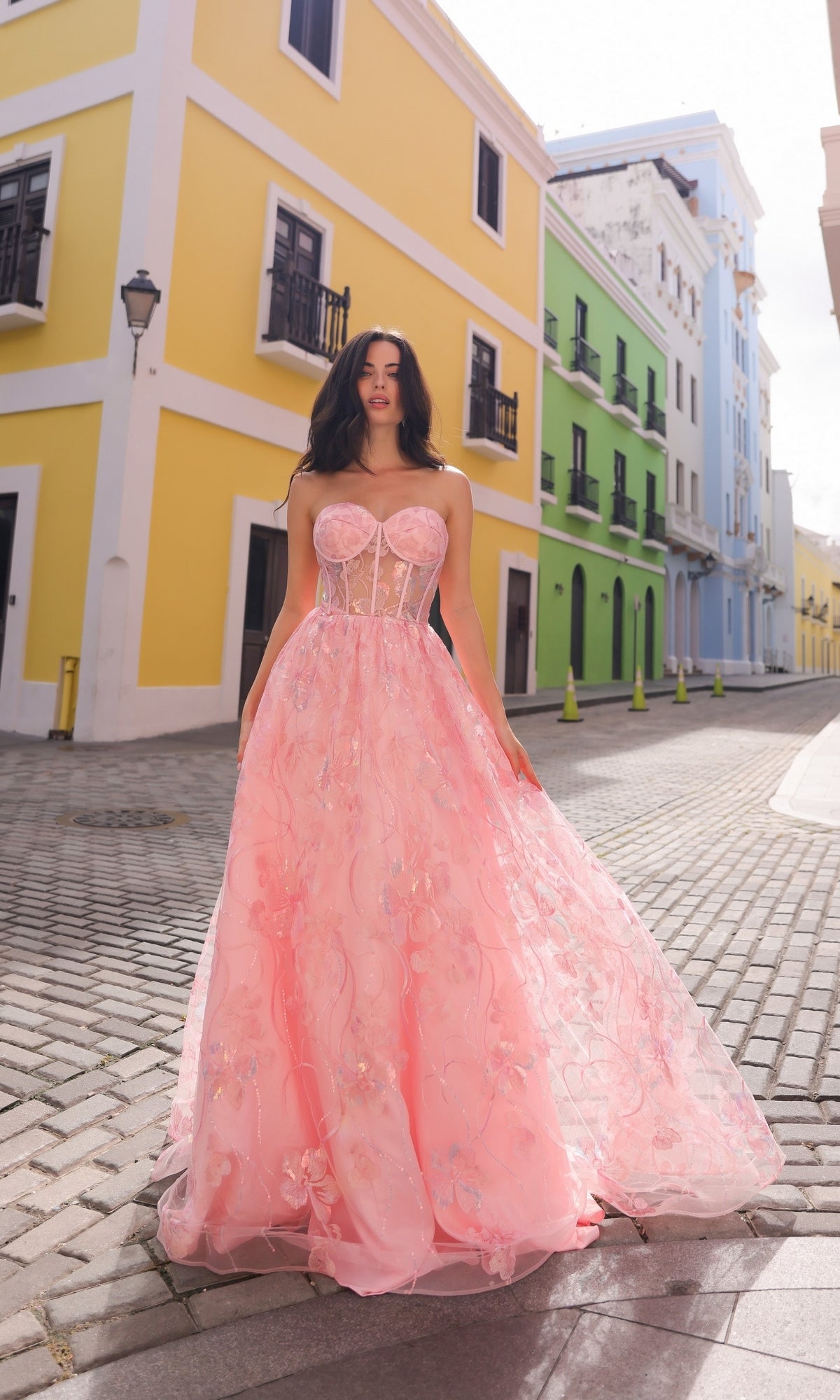 Wearing the Sequin-Floral Strapless Long Prom Ball Gown T1332, a woman stands on a cobblestone street lined with vibrant yellow, green, and blue buildings.