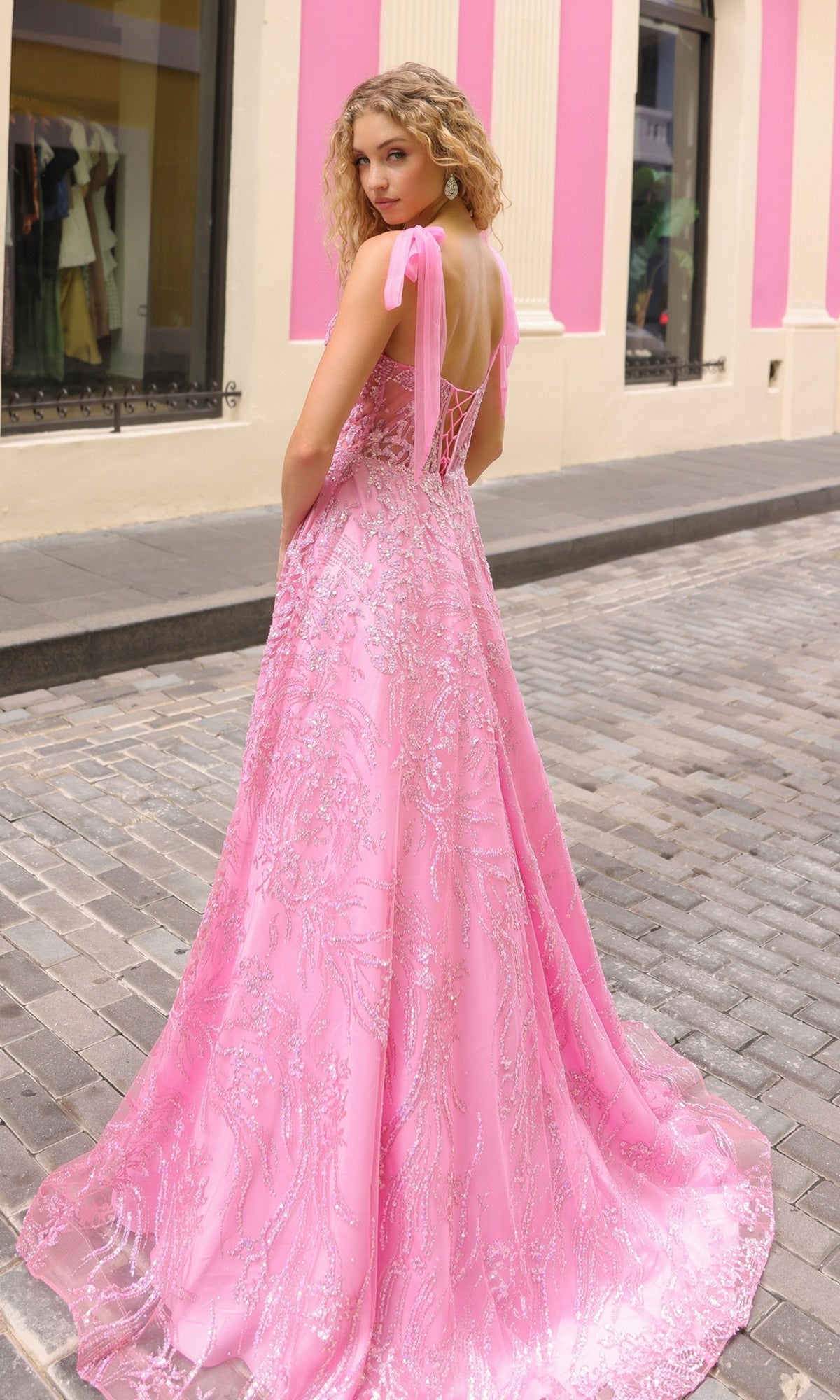 A woman with curly blonde hair wears the Sequin Long Prom Ball Gown with Shoulder Bows T1336, featuring a lace-up corset bodice, as she stands on a cobblestone street in front of pink and cream walls.