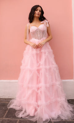 Against a pink wall, a woman wears the Shoulder-Bows Long Ruffled Prom Gown T1340—a light pink, floor-length tulle dress with ruffled layers and a fitted beaded corset bodice with delicate ribbon straps.