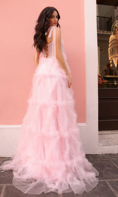 A woman in the Shoulder-Bows Long Ruffled Prom Gown T1340, a long light pink tulle dress with a beaded corset bodice and lace-up back, stands outdoors against a pale pink wall, glancing over her shoulder.