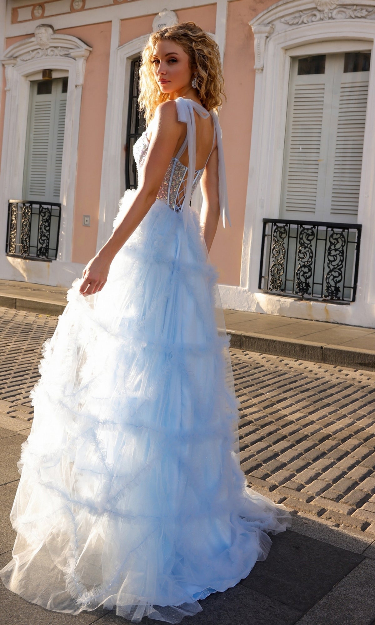 A woman in the Shoulder-Bows Long Ruffled Prom Gown T1340 stands outdoors on a sunlit street, framed by a pastel building with white shutters.
