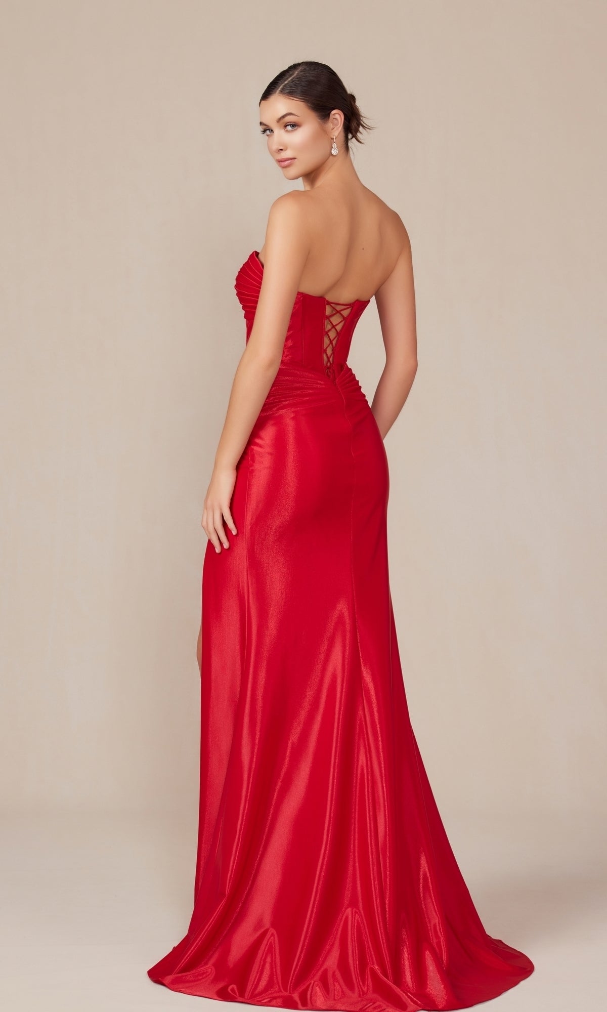 A woman wearing the Nox Anabel T1489 Strapless Lace-Up Long Prom Gown, a floor-length red satin dress with a lace-up back, poses looking over her shoulder against a neutral background.