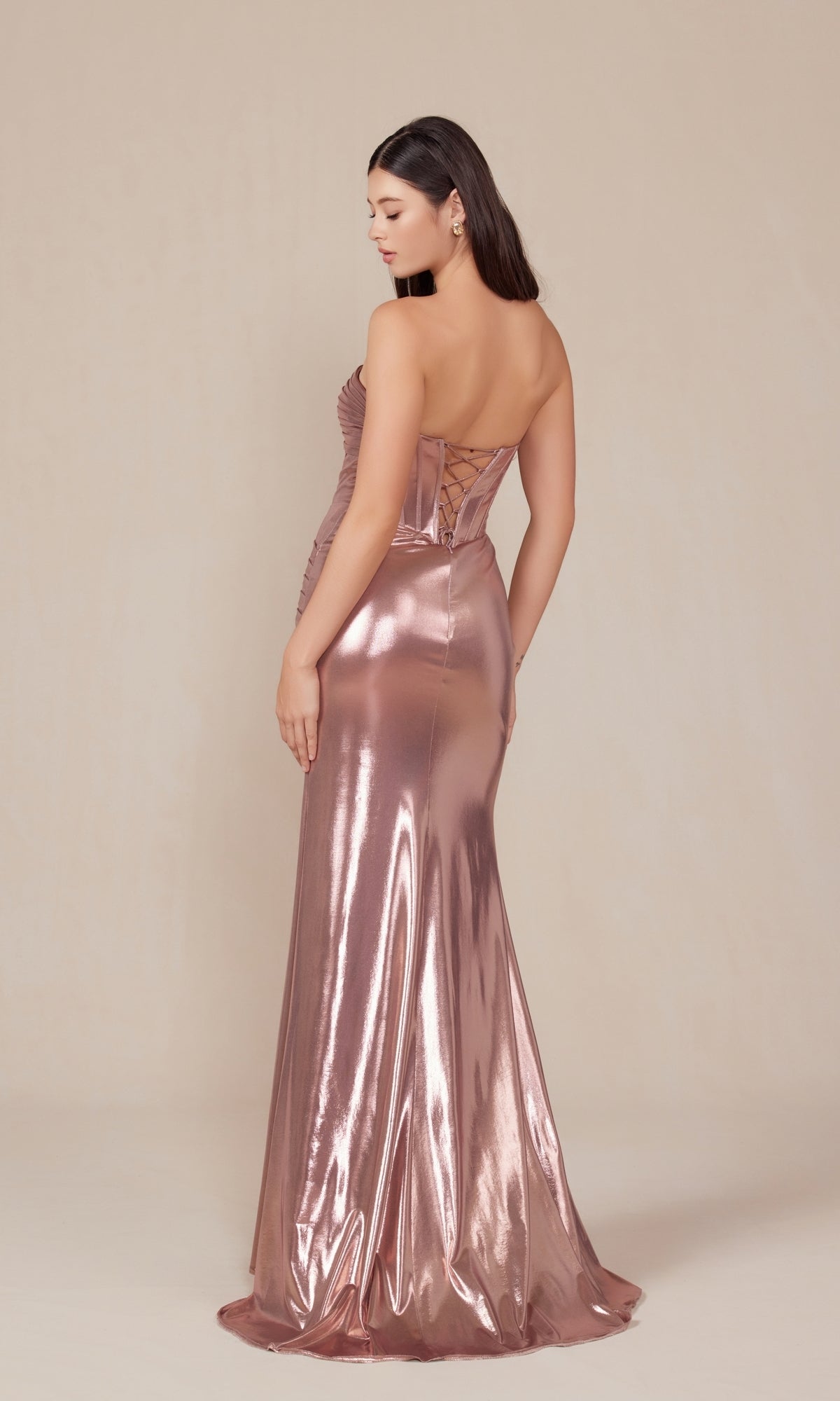 A woman faces away, wearing the Pleated Metallic Long Prom Dress Nox Anabel T1499 in rose gold, featuring a strapless neckline, lace-up corset back, fitted floor-length silhouette, and pleated details against a plain background.