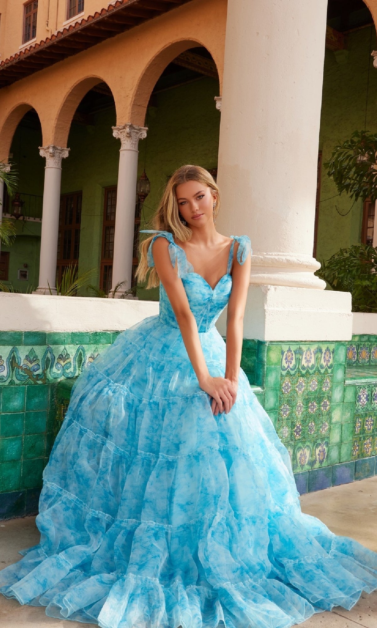 A woman wears the Nox Anabel T1501 Tie-Dye Long Ruffle Prom Dress, posing before a tiled wall with columns and an arched building in the background.