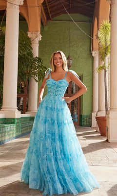 A woman stands outdoors by columns and greenery, wearing the Nox Anabel T1501 Tie-Dye Long Ruffle Prom Dress featuring shoulder ties and a sweetheart neckline.