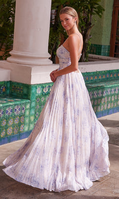 A woman stands outdoors by a tiled wall and pillar in the Nox Anabel T1506 long prom dress, featuring thin straps, a lace-up back, pleated A-line silhouette, and a light floral print.