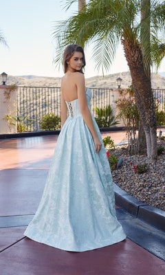 A woman wearing the Nox Anabel T1912 Long Prom Dress, featuring a strapless lace-up back and light blue floral jacquard fabric, stands outdoors by palm trees with her back to the camera.