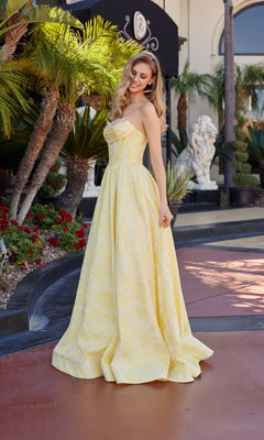 A woman wearing the Nox Anabel T1912 long prom dress with a jeweled sweetheart neckline stands outdoors by palm trees and red flowers, looking down and smiling.