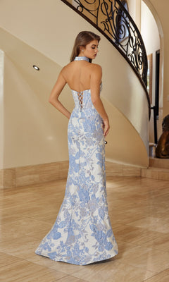 A woman stands indoors by an ornate staircase, showcasing the stunning backless design of the Nox Anabel T1922 long prom dress—a floor-length blue and white floral brocade gown with a corset bodice and lace-up detail.