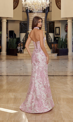 A woman stands indoors on marble flooring, facing away, wearing the Nox Anabel T1922 long pink and white floral brocade prom dress with a corset bodice. A grand staircase and chandelier are in the background.