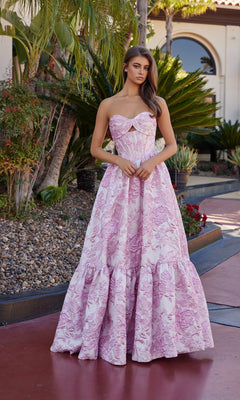 A woman stands outside in front of plants and a building, wearing the Nox Anabel T1923 long pink prom dress with a strapless, floor-length silhouette and subtle sweetheart neckline.