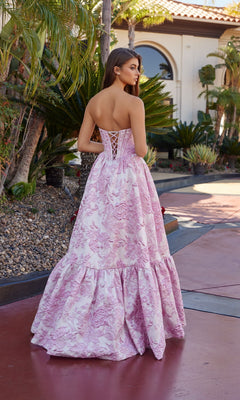 A woman stands outdoors with her hair down, wearing the Nox Anabel T1923 long prom dress—strapless, with a corset bodice, lace-up back, and delicate pink floral brocade patterns.