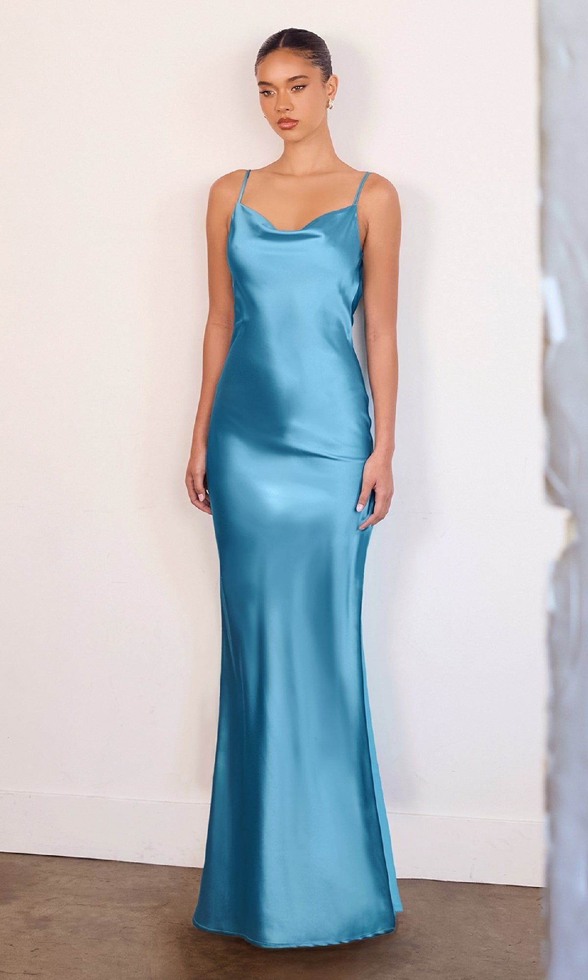 A woman poses against a plain wall in the Astee T2000 Cowl-Back Sexy Long Prom Dress, a fitted, floor-length satin gown with thin straps in light blue.